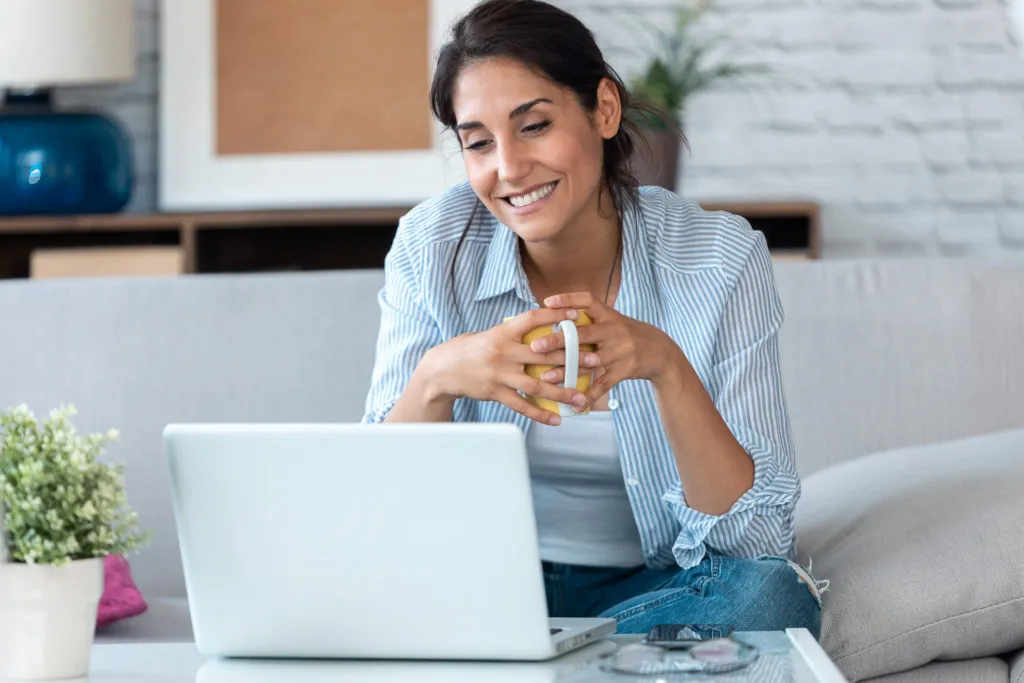 happy business woman working remotely from home with laptop and drinking coffee while sitting on sofa.